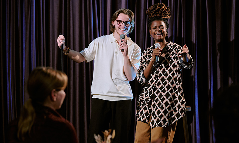 Deux personnes font face au public sur scène, des micros à la main, pendant un spectacle de stand-up